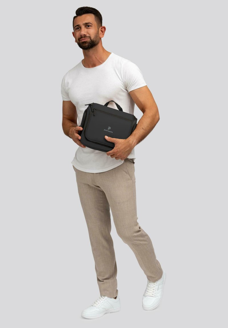 Bolso tipo mensajero sintético negro con cierre de cremallera, asa superior y detalle del logo, sostenido por una persona que lleva una camisa blanca ajustada y pantalones claros.