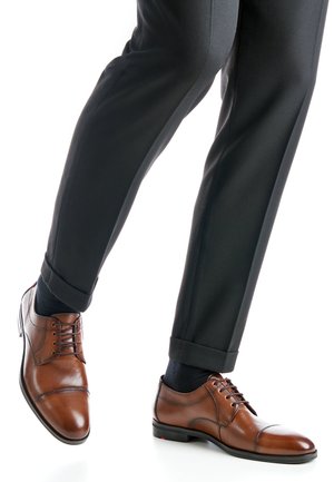 Brown leather oxford shoes with perforated design, lace-up closure, and contrasting textured accents, paired with black tailored trousers.