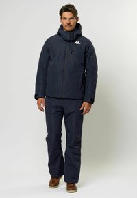 Mann in einer marineblauen Winterjacke und -hose mit weißem Logo, stehend vor einem schlichten hellen Hintergrund, braune Stiefel sichtbar.