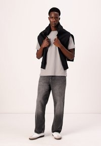 Grå bomulds t-shirt og mørkegrå jeans, med en sort sweater draperet over skuldrene. Hvide sneakers tilføjer kontrast til outfittet.