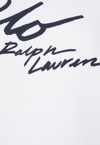 Signature cursive noire "Ralph Lauren" sur un fond en tissu blanc, probablement un gros plan d'un vêtement de marque.
