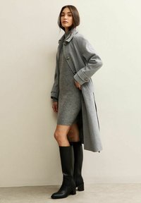 Cappotto di lana grigio con un ampio colletto e bottoni, indossato sopra un vestito fitted di maglia grigia, abbinato a stivali alti in pelle nera. Texture lisce.