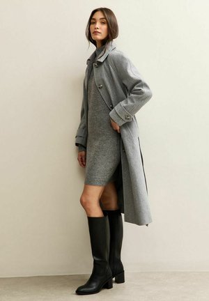 Abrigo de lana gris con un amplio cuello y botones, superpuesto a un vestido de punto gris ajustado, combinado con altas botas de cuero negras. Texturas suaves.