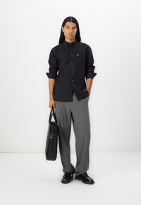 Camicia nera a maniche lunghe con taschino e logo giallo, abbinata a pantaloni grigi a quadri e con una borsa a tracolla nera.