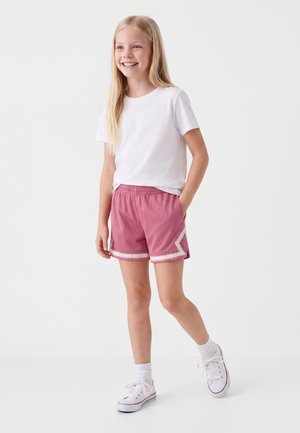 Niña joven con cabello rubio largo que lleva una camiseta blanca, pantalones cortos rosas con ribete blanco, calcetines blancos y zapatillas blancas, sonriendo y posando.