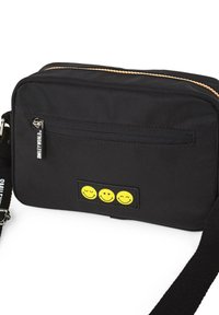 Bolso cruzado de tela negra con un bolsillo con cremallera, acentos de carita feliz amarilla y una correa ajustable. Material texturizado con cremallera en contraste.