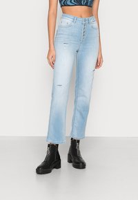 Jeans de cintura alta, azul claro, com corte reto, apresentando detalhes desgastados e fecho com botão na frente. Combinados com botas de tornozelo pretas.