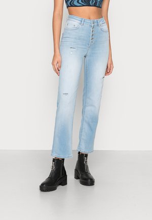 Jeans de cintura alta, azul claro, com corte reto, apresentando detalhes desgastados e fecho com botão na frente. Combinados com botas de tornozelo pretas.