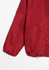 Manche de veste légère rouge avec poignet élastique et grande poche diagonale, présentée sur fond blanc.