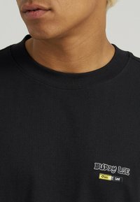 Sort bomulds sweatshirt med rund halsudskæring, der prydes af det broderede "BUDDY LEE" logo og et lille gult mærke på venstre bryst. Glat tekstur.