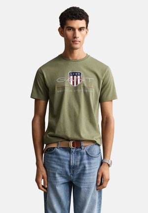 Jeune homme aux cheveux courts et bouclés portant un T-shirt olive avec le logo GANT, un jean bleu clair, une ceinture beige et une montre-bracelet, debout sur un fond blanc.