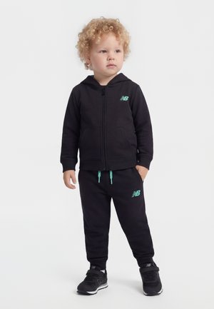 Bambino piccolo con capelli biondi ricci, in piedi, indossa una felpa nera New Balance con cappuccio, pantaloni della tuta coordinati e scarpe da ginnastica nere su uno sfondo bianco.