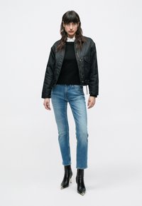 Μαύρο σατινέ μπουφάν bomber, μαύρο πλεκτό πουλόβερ, μπλε τζιν και μαύρα μποτάκια αστραγάλου με χρυσές λεπτομέρειες. Το μοντέλο στέκεται μπροστά σε απλό φόντο.