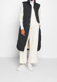Person som bär en lång svart vadderad väst över en vit polotröja, breda krämfärgade byxor och vita plattforms-sneakers, stående mot en vit vägg.