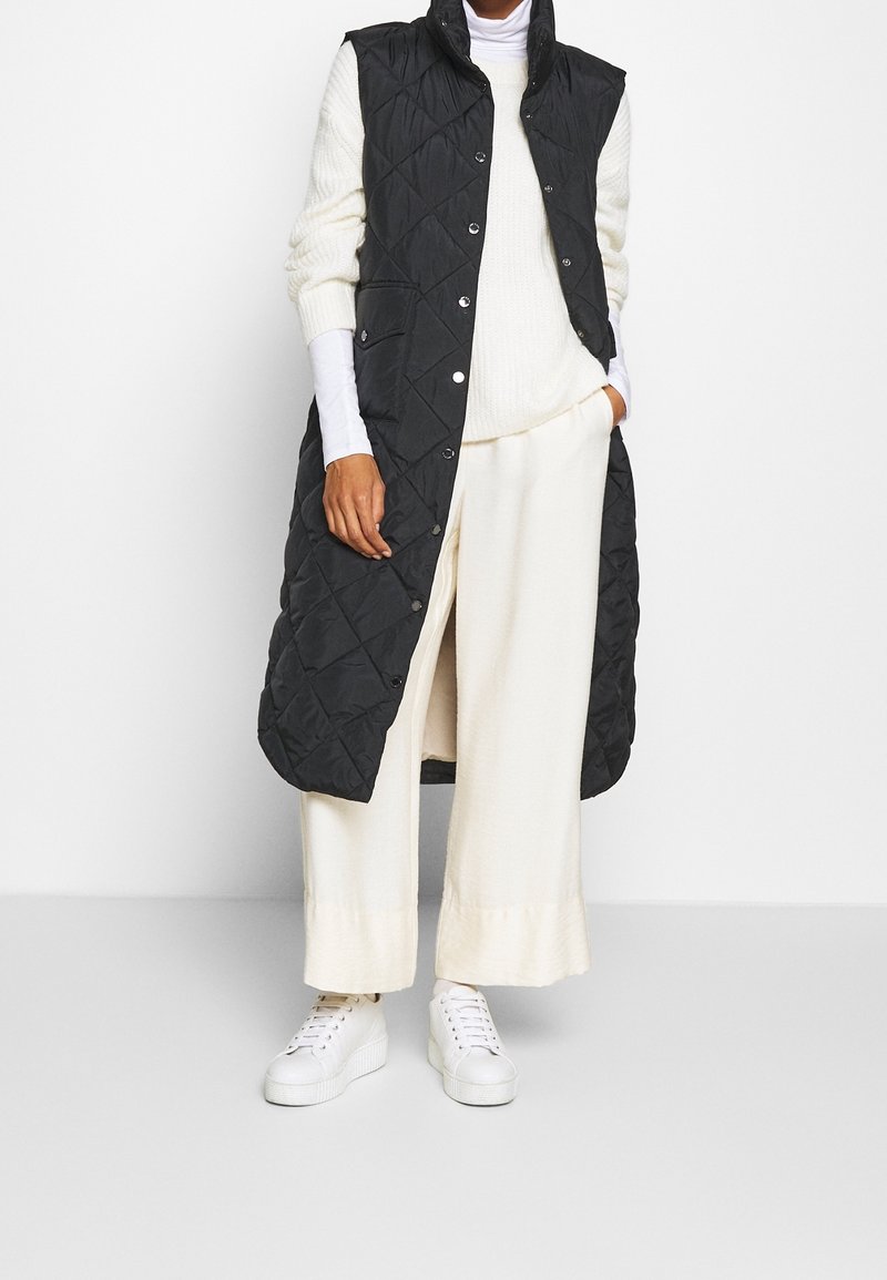 Person som bär en lång svart vadderad väst över en vit polotröja, breda krämfärgade byxor och vita plattforms-sneakers, stående mot en vit vägg.