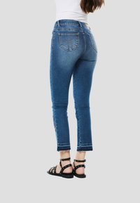 Modré džínsové nohavice s priliehavým strihom, skrátenou dĺžkou, ošúchanými lemami a ozdobnými detailmi na ľavej strane; v kombinácii s čiernymi remienkovými sandálmi.