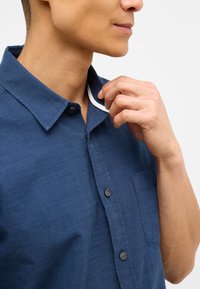 Navy blue koszula zapinana na guziki z fakturowanej tkaniny, z kołnierzykiem w kształcie szpilki, kieszonką na piersi i kontrastującym wykończeniem wewnętrznego kołnierzyka.