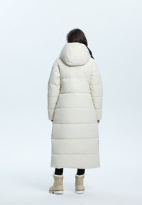 Lange, puffy, licht crèmekleurige jas met een capuchon, met een doorgestikt ontwerp en ribbelmanchetten, gedragen over zwarte broeken en tan laarzen.