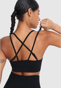 Žena s pletenými vlasmi upravujúca čierny popruh na svojom športovom podprsenke, pričom ukazuje tetovanie na svojom hornom ramene.