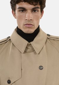 Gabardina beige con gran cuello, diseño de doble botonadura y detalles en negro. Llevada sobre un suéter de cuello alto negro. Tejido texturizado.