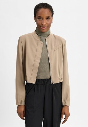 Frau mit kurzen lockigen Haaren, die kleine Creolen trägt, eine beige cropped Jacke über einem schimmernden goldenen Rollkragenpullover und hochgeschnittene schwarze Hosen.