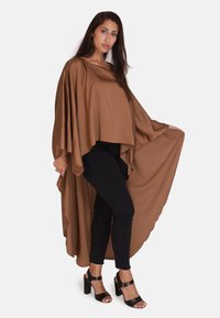 Top marrón fluido de doble capa con mangas anchas y un dobladillo asimétrico. Combinado con jeans ajustados negros y tacones abiertos negros.
