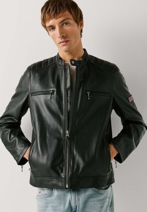 Ung mand iført en sort læderjakke med lynlåslommer og et Union Jack-patch på ærmet, hænderne i lommerne, stående op ad en hvid væg.