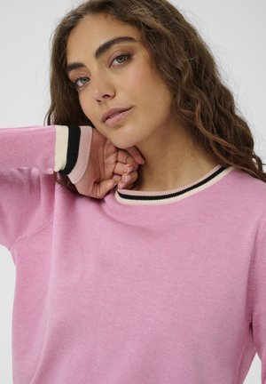 Rózsaszín hosszú ujjú pulóver, bordázott fekete és krémszínű gallérral, puha tapintású, laza szabású. Közeli kép a nyakvonalról és a testtartásról.