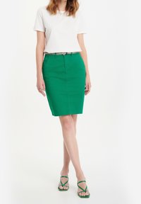 Bílý krátký rukáv top s ozdobami, spojený se zelenou tužkovou sukní a zelenými sandály na pásku; elegantní, přiléhavý design.