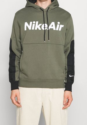 Osoba ubrana w oliwkową bluzę z kapturem Nike Air z czarnymi łatami na łokciach i kremowe spodnie, stojąca na jednolitym tle.