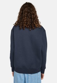 Felpa blu navy con vestibilità rilassata, polsini e orlo a costine. Tessuto liscio, senza motivi o accenti visibili. Indossata con pantaloni azzurri.