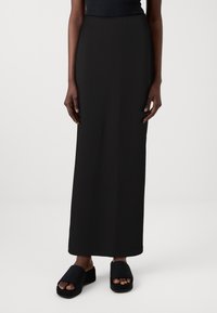 Schwarzer gerippter Maxirock mit gerader Silhouette, kombiniert mit schwarzen Plateau-Sandalen. Glatte Textur, keine sichtbaren Muster oder Beschläge.