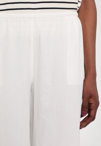 Main droite avec une peau foncée et du vernis à ongles violet à côté d'un pantalon blanc à taille élastique et d'un haut rayé noir et blanc.