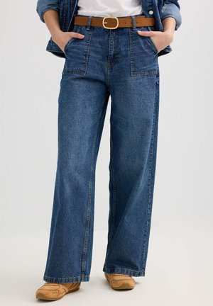 Persona con jeans azules de talle alto y pierna ancha, con grandes bolsillos frontales, cinturón marrón, chaqueta de mezclilla, camiseta blanca y zapatos casuales color crema.