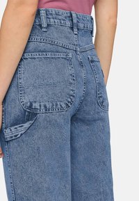 Mulher a usar um par de jeans de ganga azuis de cintura alta com grandes bolsos traseiros e um pequeno laço lateral, combinados com um top rosa.