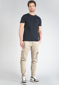 Zwarte T-shirt met korte mouwen en een klein logo, gecombineerd met beige cargobroek met meerdere ritszakken, en zwart-witte sneakers.