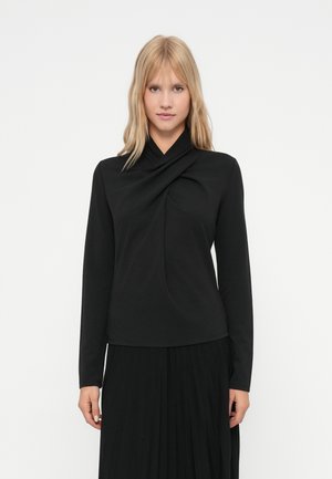 Mlada ženska z blond lasmi nosi črn zgornji del z dolgimi rokavi z drapiranim izrezom in črno plisirano krilo na bež ozadju.