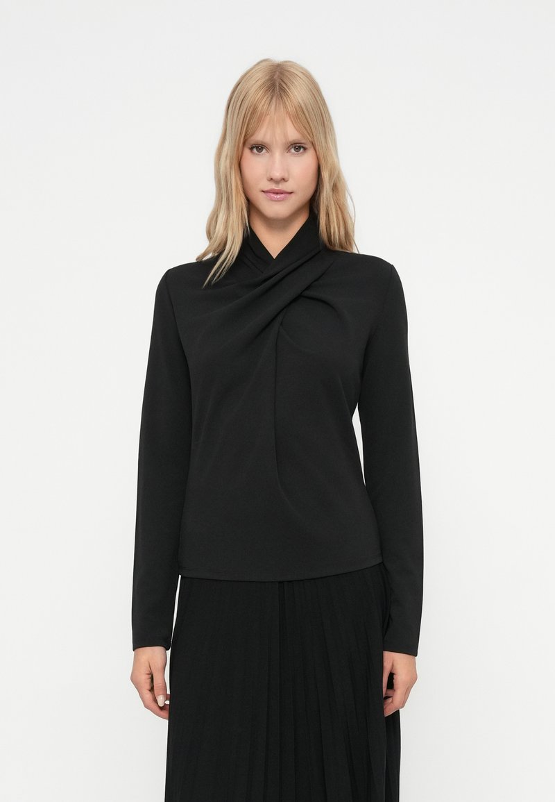 Jauna sieviete ar blondiem matiem valkā melnu garām piedurknēm topu ar drapētu dekoltē un melnu svārku ar krokām uz baltas fona.