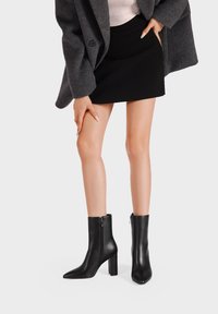 Stivali neri in pelle fino alla caviglia con punta appuntita e tacco grosso. Presentano una zip laterale e una texture liscia. Indossati con una gonna nera.