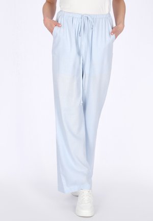 Persoon die lichtblauwe wijde broek met trekkoord draagt, met handen in de zakken en witte sneakers, staand tegen een witte achtergrond.