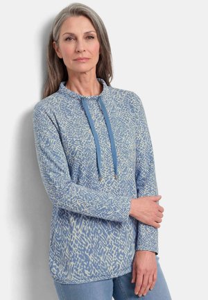 Eine Frau mittleren Alters mit langen grauen Haaren trägt ein blau gemustertes Langarmshirt und blaue Jeans und steht vor einem hellen Hintergrund.