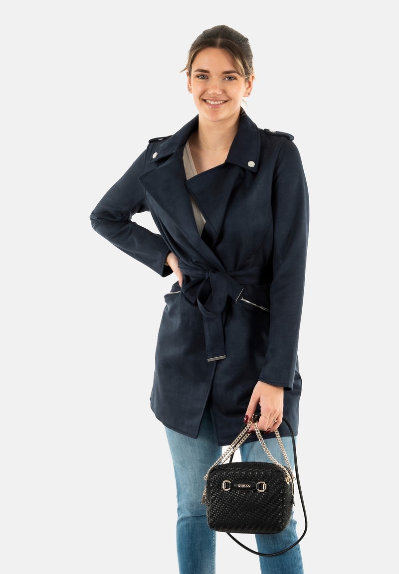 Trench coat blu navy con cintura, accessori in argento e vita cintrata. Abbinato a una piccola borsa nera, esposto su uno sfondo bianco.