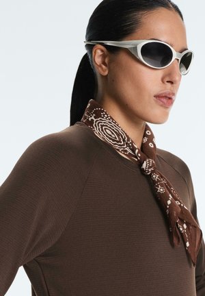 Nő hátracsíkozott hajjal, futurisztikus fehér napszemüveget, barna mintás nyakbékítőt és sötétbarna felsőt visel, jobbra néz.