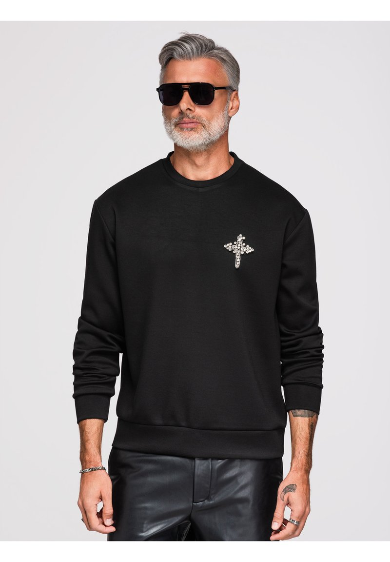 Homme aux cheveux gris et à la barbe portant des lunettes de soleil noires, un sweat-shirt noir avec une croix ornée de bijoux, et un pantalon en cuir noir sur fond uni.