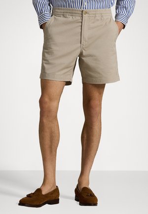 Beige katoenen shorts met een elastische tailleband, twee zijzakken en een knoopsluiting. Draagt daarbij bruine suède instapschoenen.