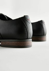 Czarne skórzane buty z teksturowanym obcasem, z brązową warstwową podeszwą. Prosty design z czerwonym akcentem w pobliżu obcasa.