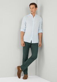 Camicia azzurro chiaro con bottoni, maniche arrotolate, abbinata a pantaloni verde scuro e scarpe in camoscio marroni. Design semplice, senza motivi o accenti visibili.