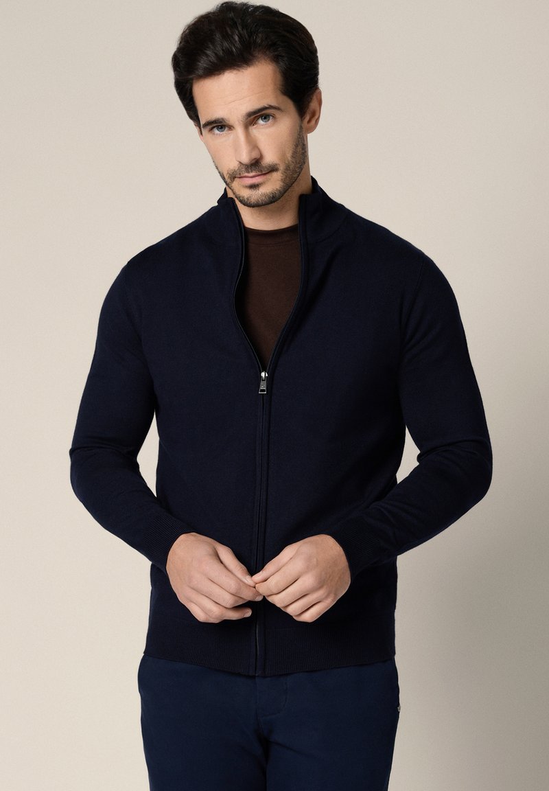 Mężczyzna z krótkimi ciemnymi włosami i brodą, ubrany w granatowy sweter na zamek błyskawiczny na ciemnej koszuli, stojący na beżowym tle.