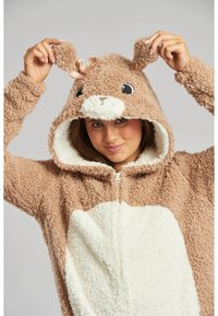 Bruine fleece onesie met een witte buik, rits aan de voorkant en een capuchon met oren en een strik. Zacht, gestructureerd stof, ideaal voor warmte.