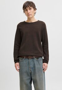 Ung person med kort brunt hår iført en mørkebrun langærmet sweater, løse blå jeans og øreringe, står foran en ensfarvet baggrund.
